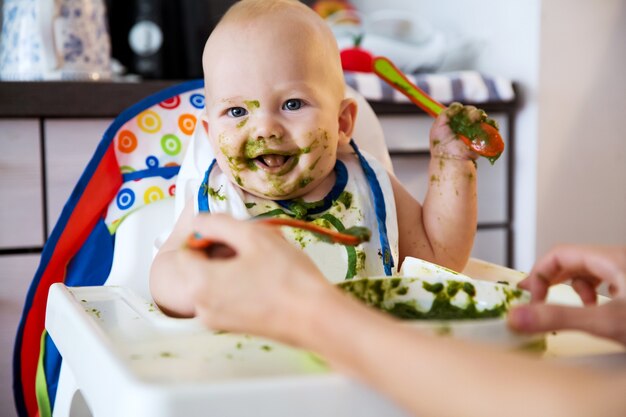 Alimentazione. Adorabile bambino che mangia con un cucchiaio nel seggiolone. Il primo cibo solido del bambino