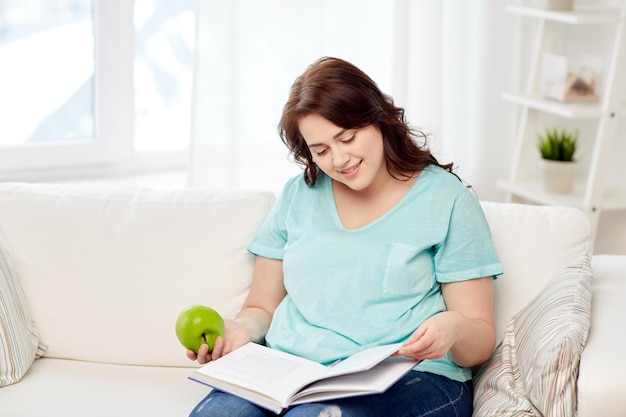 alimentazione sana, alimenti biologici, frutta, dieta e concetto di persone - felice giovane donna più taglia che legge libro e mangia mela verde a casa
