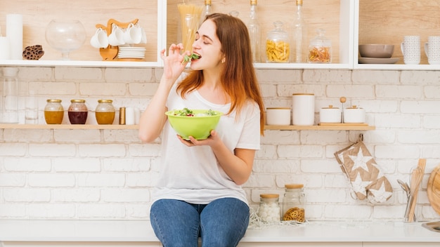 Alimentazione sana. Cibo dietetico e fitness femminile. Giovane donna che mangia insalata da una ciotola seduta su un bancone della cucina.