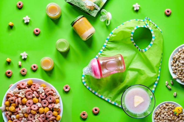 Alimenti per bambini Cereali purea di frutta e verdura cereali di grano una bottiglia per miscele e latte un ciuccio e un grembiule per bambini su sfondo verde Il concetto di alimentazione sana per i bambini