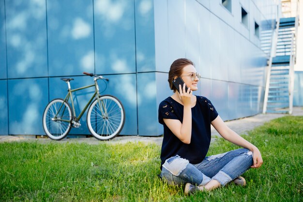 All'aperto foto di donna attraente allegra, con piacevole conversazione telefonica, appoggiato sull'erba dopo aver guidato una bici. All'aperto. Concetto di tempo libero.