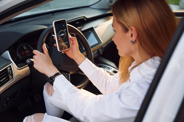 All'interno di un veicolo Con smartphone Giovane donna in abiti bianchi è con la sua auto elettrica durante il giorno