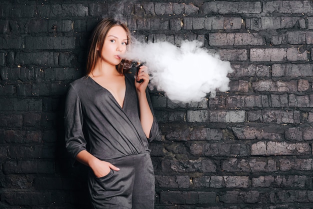 Alla moda bella ragazza che fuma una sigaretta senza tabacco, espirando grandi nuvole di fumo. Vestito con abiti alla moda.