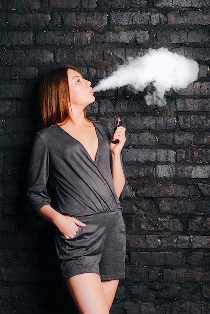 Alla moda bella ragazza che fuma una sigaretta senza tabacco, espirando grandi nuvole di fumo. Vestito con abiti alla moda.