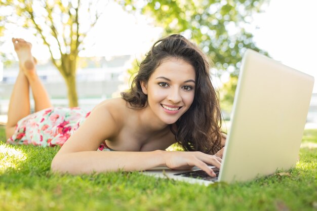 Alla moda bruna sorridente sdraiato sull&#39;erba usando il suo computer portatile