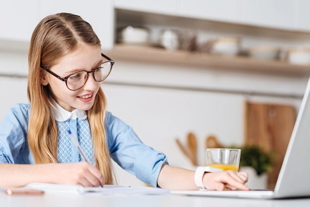 Alla ricerca di linee guida. Brillante bella ragazza positiva utilizzando il suo computer mentre si lavora sul suo compito a casa e prendere appunti