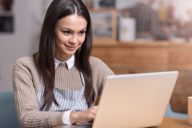 Alla ricerca di qualcosa di nuovo. Affascinante bella donna sorridente e seduta al tavolo durante l'utilizzo di laptop.