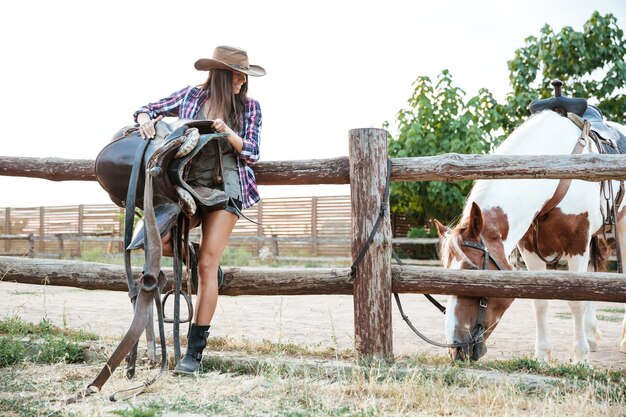 Allegra attraente giovane donna cowgirl in cappello che tiene la sella per andare a cavallo