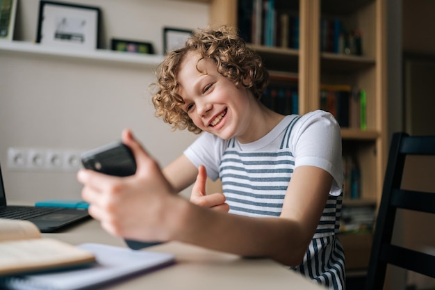 Allegra bambina riccia che tiene e utilizza il telefono cellulare mostrando il pollice in su navigando nelle app dei social network navigando in internet e scorrendo il feed di notizie