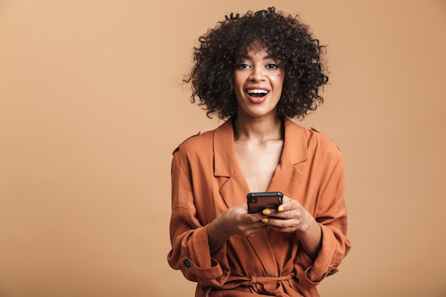 Allegra bella donna africana in possesso di smartphone e guardando direttamente su sfondo marrone