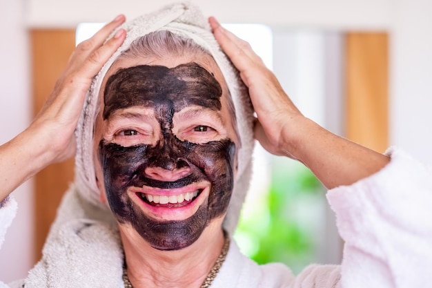 Allegra bella donna anziana in accappatoio che applica una maschera facciale detox al carbone fatta in casa guardando il benessere della fotocamera prendersi cura del concetto di pelle