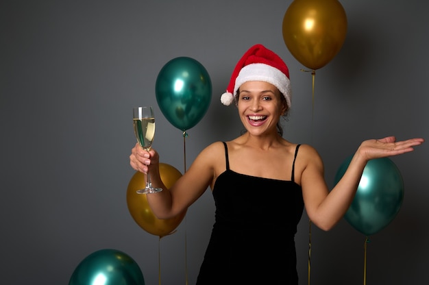 Allegra bella donna con cappello da Babbo Natale esulta e si diverte alla festa di Natale tiene un flauto di champagne e copia spazio sul palmo della mano, isolato su sfondo grigio con oro lucido e mongolfiere