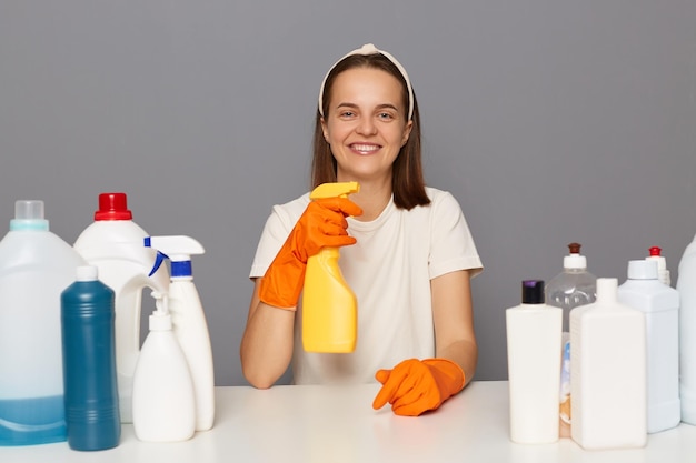 Allegra bella donna in posa sul posto di lavoro con detergenti per la pulizia isolate su uno sfondo grigio tenendo spray per la pulizia guardando la telecamera facendo le faccende domestiche