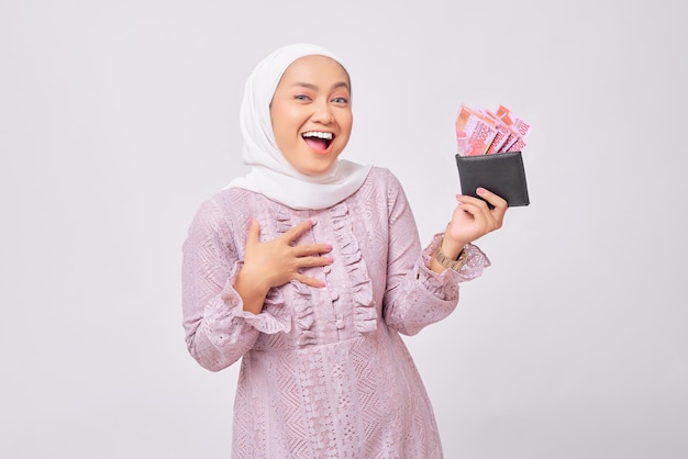 Allegra bella giovane donna musulmana asiatica che indossa hijab e abito viola che tiene portafoglio di denaro contante pieno isolato su sfondo bianco studio
