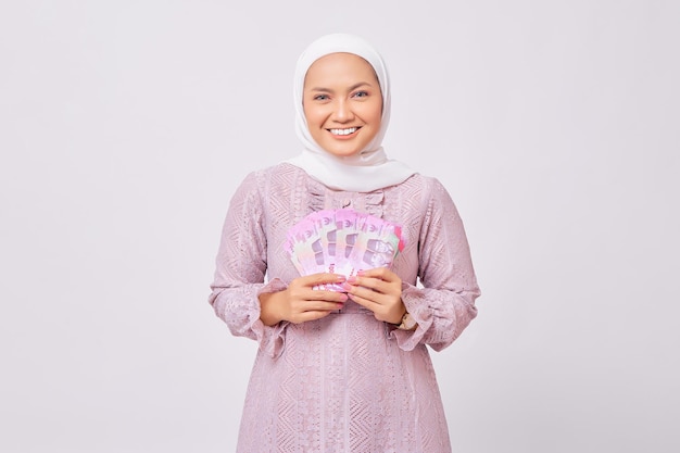 Allegra bella giovane donna musulmana asiatica che indossa l'hijab e abito viola che tiene denaro contante rupia banconote isolate su sfondo bianco studio