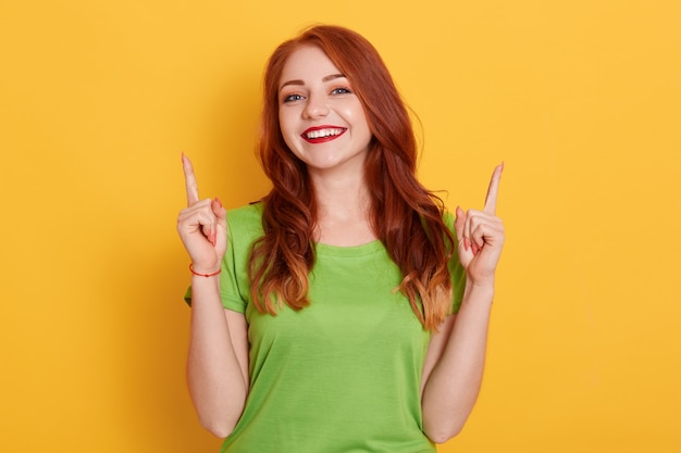 Allegra bella ragazza che punta le dita verso l'alto e guardando sorridente alla telecamera, indossa una maglietta verde, ha i capelli rossi ondulati, in piedi con lo sguardo felice contro il muro giallo.