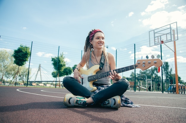 Allegra bella signora emotiva con i dreadlocks sorridendo e guardando felice mentre era seduto con la sua chitarra sul campo sportivo