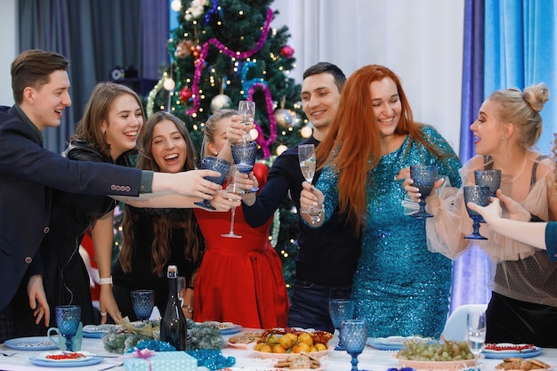 Allegra compagnia di amici festeggia il Natale o il Capodanno a un tavolo vicino all'albero di Natale con ghirlande. Le donne ridono, brindano con lo champagne.