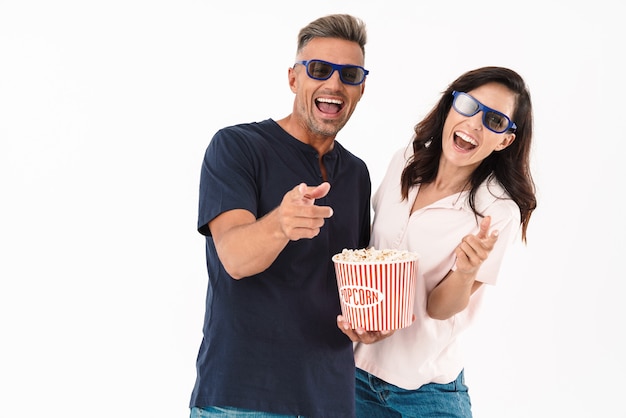 Allegra coppia attraente che indossa un abito casual in piedi isolato su un muro bianco, guardando un film con popcorn e occhiali 3d