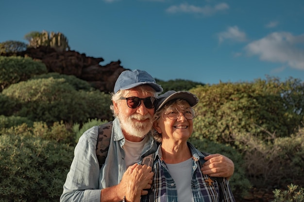 Allegra coppia caucasica anziana che si abbraccia nel trekking in montagna godendosi la natura e la libertà Vecchi pensionati sorridenti in cappello e abiti casual tra cespugli verdi e cielo blu