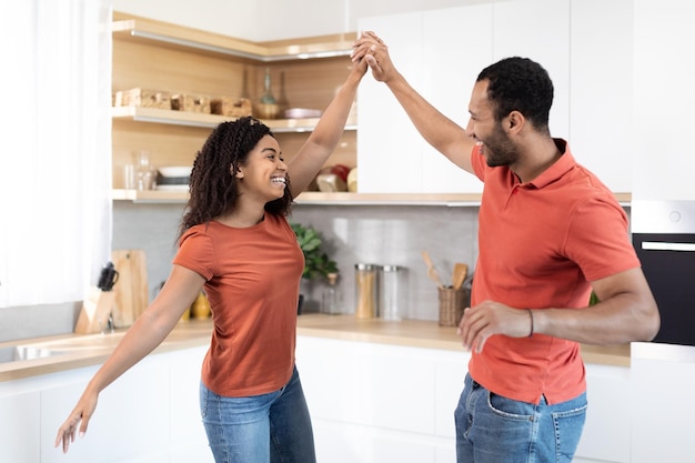 Allegra coppia nera millenaria in magliette rosse che ballano si divertono insieme alla musica nello spazio libero interno della cucina