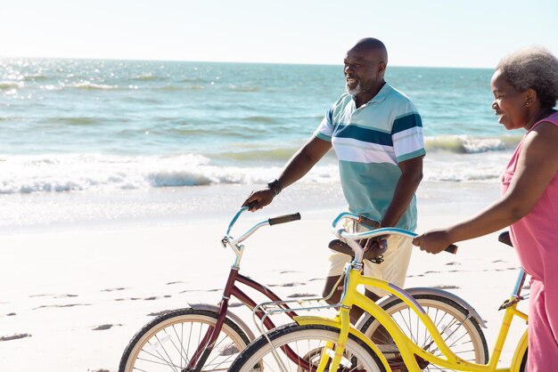 Allegra coppia senior afroamericana con le biciclette che si divertono in spiaggia contro il paesaggio marino e il cielo. Copia spazio, estate, inalterato, vacanza, amore, insieme, pensione, natura e stile di vita attivo.