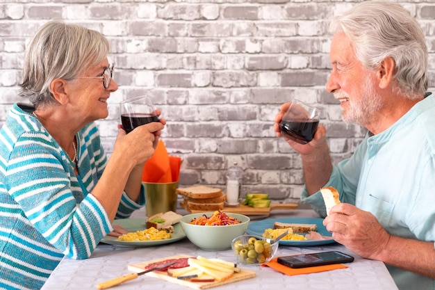 Allegra coppia senior caucasica che brinda con un bicchiere di vino rosso mentre si siede faccia a faccia a tavola con un pasto o un brunch insieme a casa