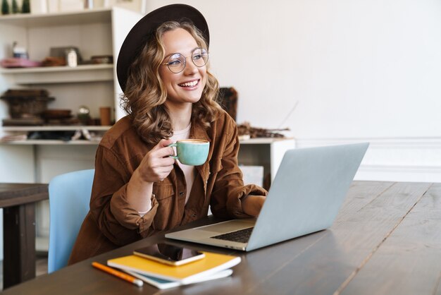 allegra donna affascinante con cappello che lavora con il computer portatile e beve caffè mentre è seduta al tavolo