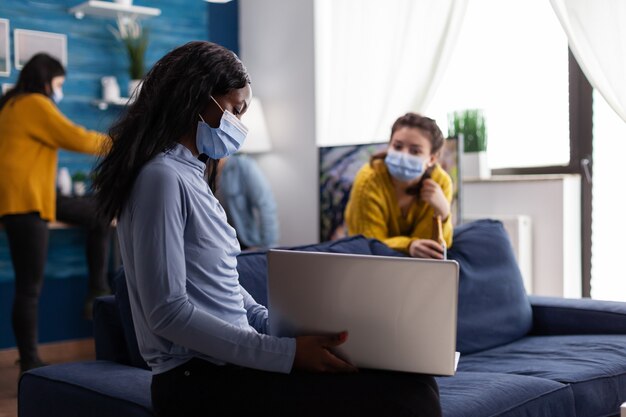 Allegra donna africana felice che mostra video sul laptop ad un amico mentre è in giro nel soggiorno dell'appartamento mantenendo le distanze sociali a causa della pandemia di coronavirus per prevenire la diffusione del virus.