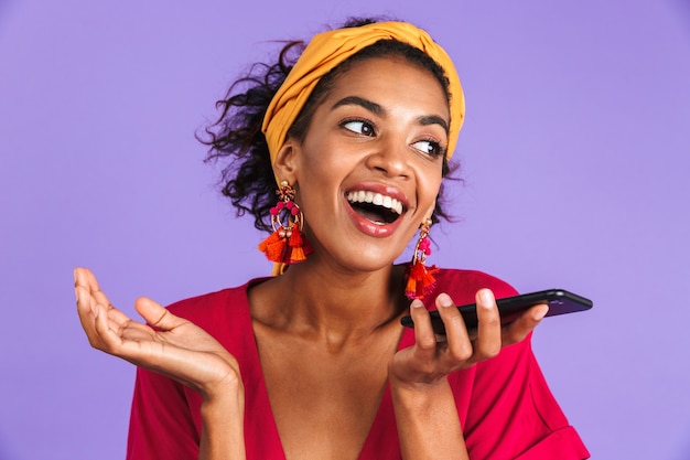 Allegra donna africana in abito parlando da smartphone e guardando lontano oltre il muro viola