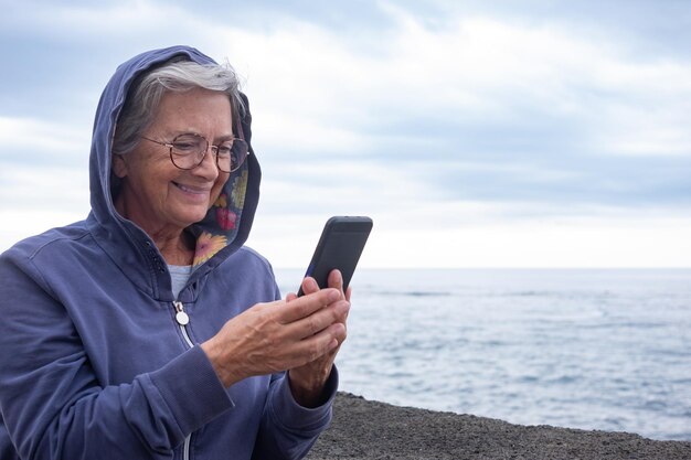 Allegra donna anziana con cappuccio sulla testa in piedi al mare in una giornata nuvolosa guardando smartphone. Donna anziana degli anni '70 che si gode la tecnologia e i social