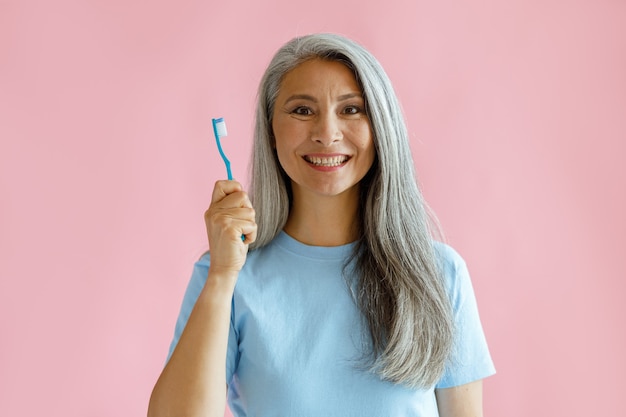 Allegra donna asiatica di mezza età con i capelli grigi in maglietta blu tiene lo spazzolino da denti in piedi su sfondo rosa in studio. Igiene del cavo orale