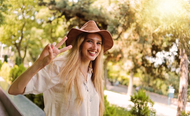 Allegra donna bionda in stile casual che indossa vestiti e cappello di feltro guarda nella telecamera e mostra il segno Piselli all'aperto