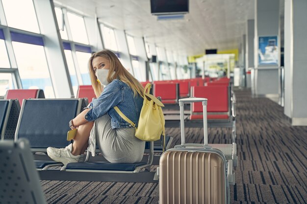 Allegra donna dai capelli lunghi che indossa una maschera protettiva mentre va in viaggio durante il periodo di quarantena