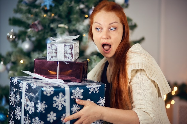 Allegra donna dai capelli rossi con un mucchio di regali per Natale vicino all'albero di Natale