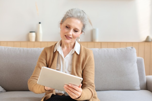 Allegra donna di mezza età seduta sul divano, utilizzando le app per tablet del computer, guardando lo schermo, leggendo buone notizie nei social network, facendo shopping o chattando online.