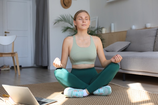 Allegra donna esile in tuta da ginnastica seduta sul pavimento a praticare yoga nella posa del loto mantenendo la calma a casa.