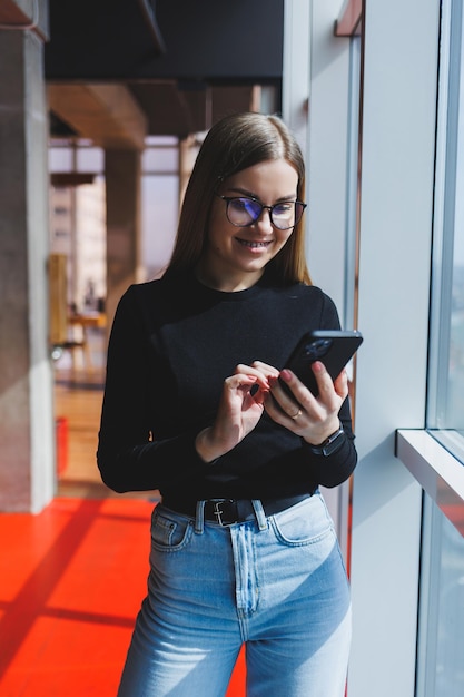 Allegra donna europea con gli occhiali che controlla la posta sui social media utilizzando smartphone e connessione wifi ragazza positiva condivide il post del blog personale mentre si gode l'app del telefono cellulare