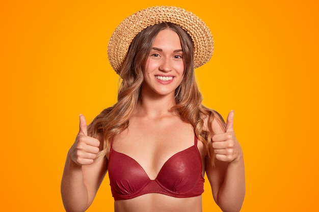 allegra donna europea con un ampio sorriso e la pelle abbronzata fa un gesto ok, indossa bikini rosso e cappello di paglia