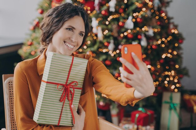 Allegra donna italiana seduta vicino al bellissimo albero di Natale a casa e prendendo selfie con un regalo