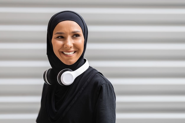 Allegra giovane atleta araba in hijab e cuffie wireless guarda lo spazio vuoto sul muro