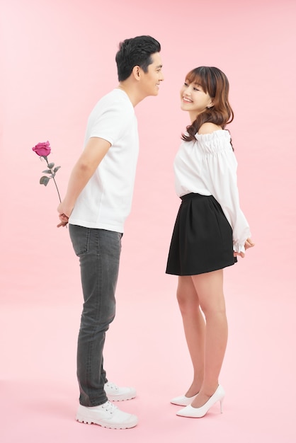 Allegra giovane coppia di innamorati che tiene una rosa e sorride mentre sta in piedi faccia a faccia e isolata su sfondo rosa