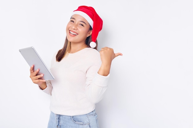 Allegra giovane donna asiatica in un cappello di Natale in possesso di una tavoletta digitale che mostra la vittoria isolata su sfondo bianco Il popolo indonesiano celebra il concetto di Natale