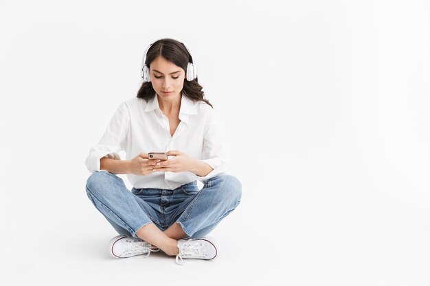 Allegra giovane donna bruna che indossa una camicia seduta isolata su un muro bianco, ascoltando musica con gli auricolari e il telefono cellulare