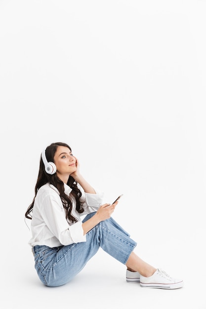 Allegra giovane donna bruna che indossa una camicia seduta isolata su un muro bianco, ascoltando musica con gli auricolari e il telefono cellulare