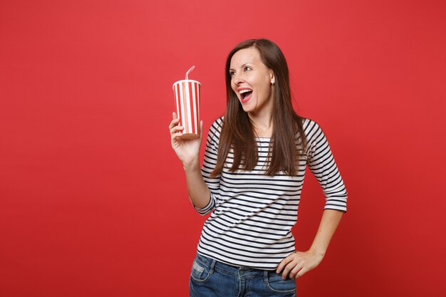 Allegra giovane donna che tiene una tazza di plastica di cola o soda, guardando da parte, tenendo la bocca spalancata isolata su sfondo rosso brillante della parete. Persone sincere emozioni, concetto di stile di vita. Mock up copia spazio.