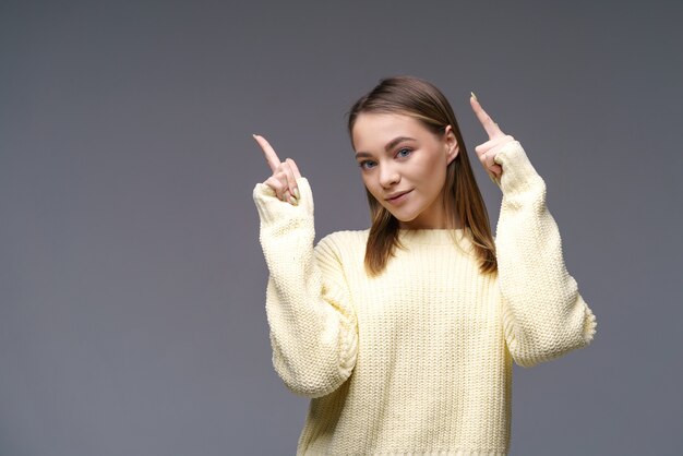 Allegra giovane donna di etnia caucasica mostra i pollici in un maglione giallo su una superficie grigia pubblicità positiva bella ragazza in posa in studio