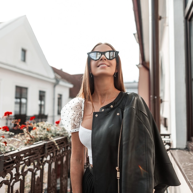 Allegra giovane donna hipster con un sorriso positivo in occhiali da sole alla moda in una giacca nera gode di relax in piedi su un balcone vintage in una giornata di sole autunnale. Ragazza allegra che riposa all'aperto.