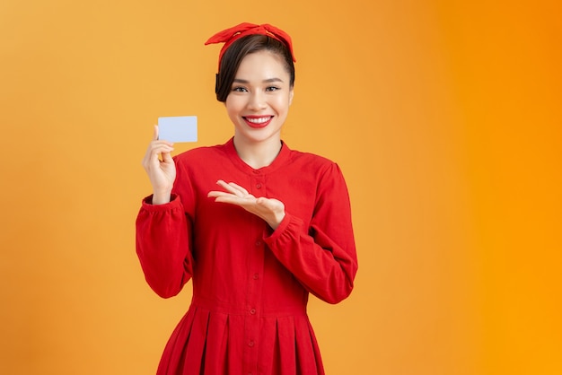 Allegra giovane donna in abito rosso in possesso di carta di credito bancaria isolata su uno sfondo arancione