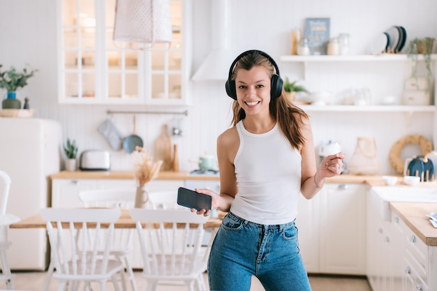 Allegra giovane donna ispanica in casual usando le cuffie e il telefono che balla in cucina guarda la fotocamera sorrisi a trentadue denti felici dalla nuova casa Bella studentessa che si diverte a casa